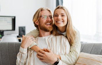 Young couple embraces on their cozy sofa at home, both smiling and radiating love and happiness in their modern living room. Relationship concept